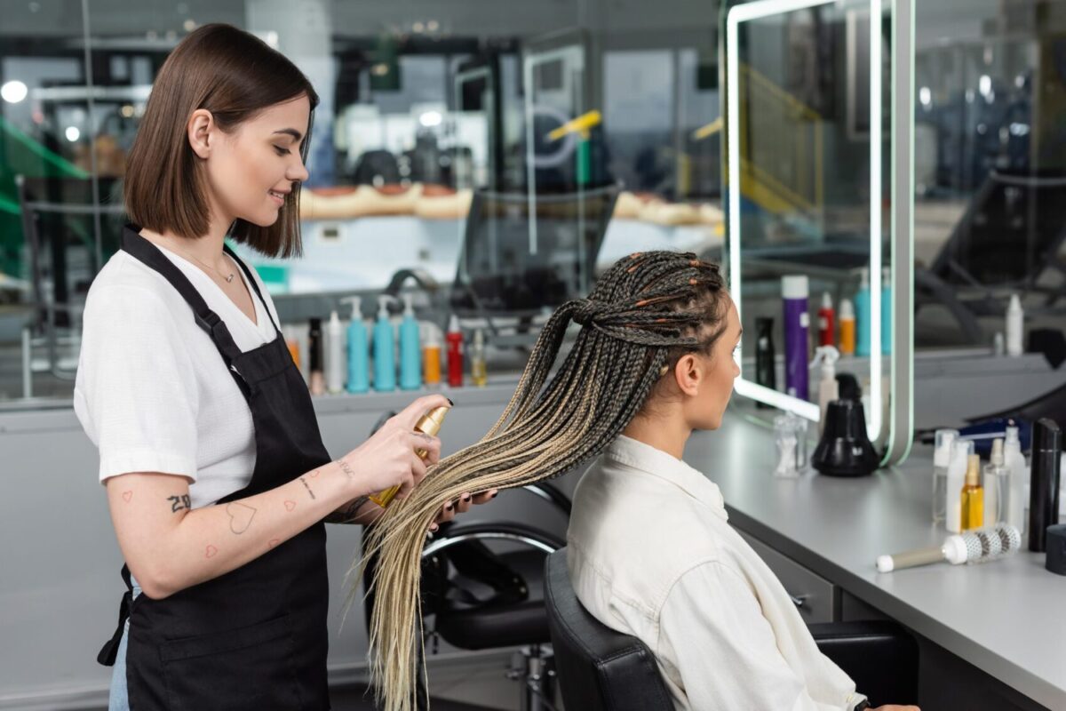 hair salon tattooed beauty worker in apron applying hair product on braids of woman in salon spray e1741071522431